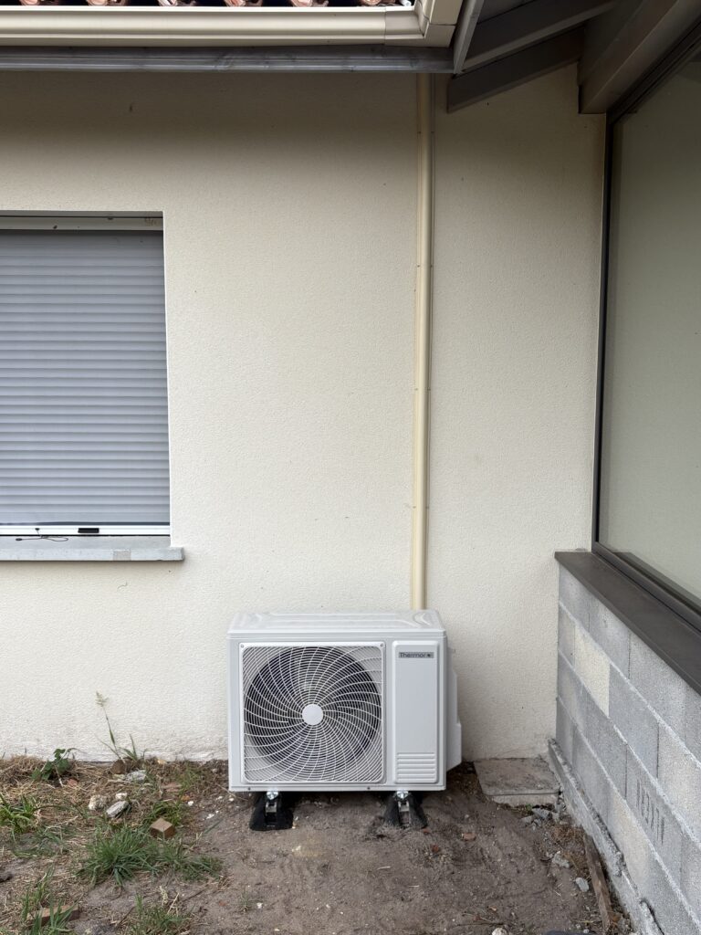 Unité extérieure de climatisation Thermor blanche installée sur le sol près d'une maison beige avec volet roulant fermé et gouttière.